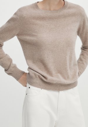 Personne portant un pull beige col rond et un pantalon blanc, les mains posées sur les hanches, sur un fond uni.