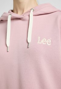 Lichtroze hoodie met witte trekkoorden, met een zachte textuur en geborduurd "Lee" logo op de borst. Zilveren metalen aglets accentueren de trekkoorden.