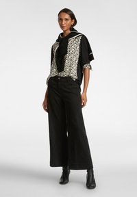 Schwarze weite Hosen kombiniert mit einem gemusterten Oberteil, das schwarze und cremefarbene Rautendesigns zeigt. Das Model trägt einen schwarzen Pullover, der am Hals gebunden ist.