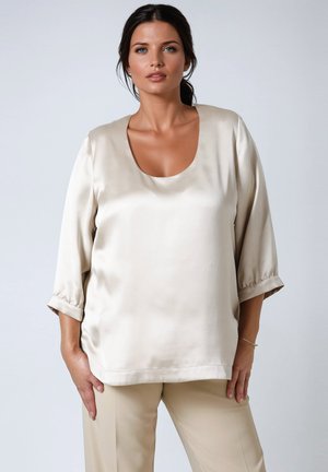 Femme aux cheveux bruns en queue de cheval basse portant un chemisier en satin crème à manches trois-quarts et un pantalon beige, se tenant devant un fond gris.