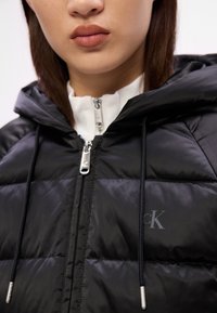 Czarna kurtka puchowa z zamkiem błyskawicznym z przodu, srebrnymi detalami i kapturem. Posiada logo CK na klatce piersiowej oraz regulowane sznurki.