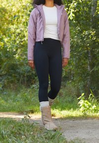 Ljuslila cropped jacka, vit t-shirt, svarta leggings, beige stövlar med vita manschetter, utomhusmiljö med grönska i bakgrunden.