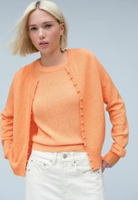 Cardigan en tricot orange avec un tissu texturé, présentant un devant boutonné et un col arrondi, associé à un pull orange assorti et un pantalon en jean blanc.