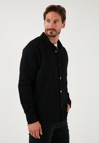 Camisa negra de botones en tejido grueso, con cuello, bolsillo en la parte delantera y herrajes plateados. Combinada con jeans oscuros.