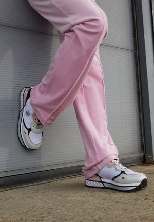 Person trägt weiße und pinke Sneakers und lockere pinke Hose, steht mit einem Fuß auf dem Boden und dem anderen an eine Wand gelehnt.