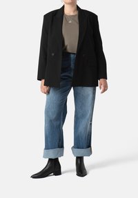 Blazer negro a medida sobre una blusa verde oliva, combinado con jeans de tiro alto de lavado claro, remangados en el dobladillo, y botas negras de tobillo.