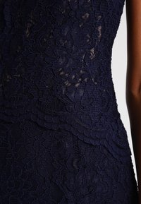 Robe en dentelle bleu marine avec des motifs floraux complexes, tissu texturé et silhouette ajustée. Présente un ourlet festonné.