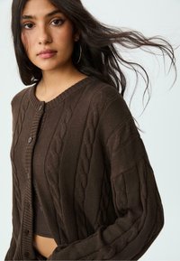 Brauner Kabelstrick-Cardigan mit rundem Ausschnitt, Knopfverschluss und langen Ärmeln, der strukturierte Muster und gerippte Akzente zeigt.