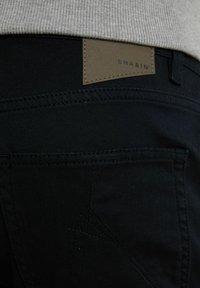 Los pantalones de mezclilla negra cuentan con una cinturilla texturizada y una etiqueta de cuero marrón grabada con "CHASIN". Se aprecian detalles de costura sutil y bolsillos traseros visibles.