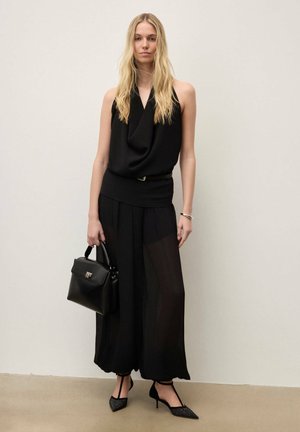 Femeie cu păr blond lung, purtând o rochie neagră fără mâneci, ținând o poșetă neagră și având pantofi negri cu vârf ascuțit, în fața unui perete simplu.