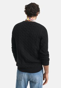 Pulover negru cu tricotaj în formă de cabluri, cu guler rotund, mansete și tiv cu dantelă. Partea din spate are un model texturat. Purtați cu blugi de culoare albastru deschis.