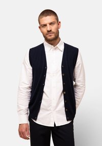 Marinblå stickad väst med knappslå fram, lager på lager över en vit skjorta, med V-halsringning och utan ärmar, i ett lätt tyg.