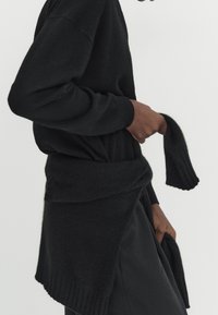 Black sweater made of soft knit material, featuring a ribbed hem and long sleeves, with a wrapped detail at the waist.