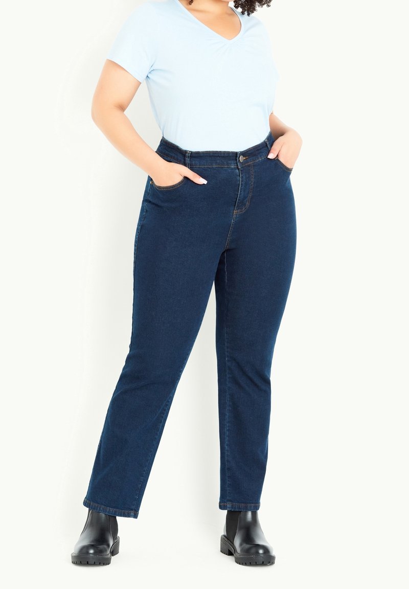 Femme debout, les mains dans les poches, portant un t-shirt bleu clair à col en V, un jean bleu foncé et des bottines noires sur fond blanc.