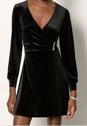 Black velvet long-sleeve mini dress with V-neck, side ruching secured by three sparkly buttons, worn with a pearl necklace and bracelet.