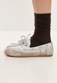 Pantofi slip-on de culoare deschisă, cu un finisaj texturat, având un detaliu cu fundiță și accente metalice, asortați cu șosete întunecate, cu densitate ridicată.