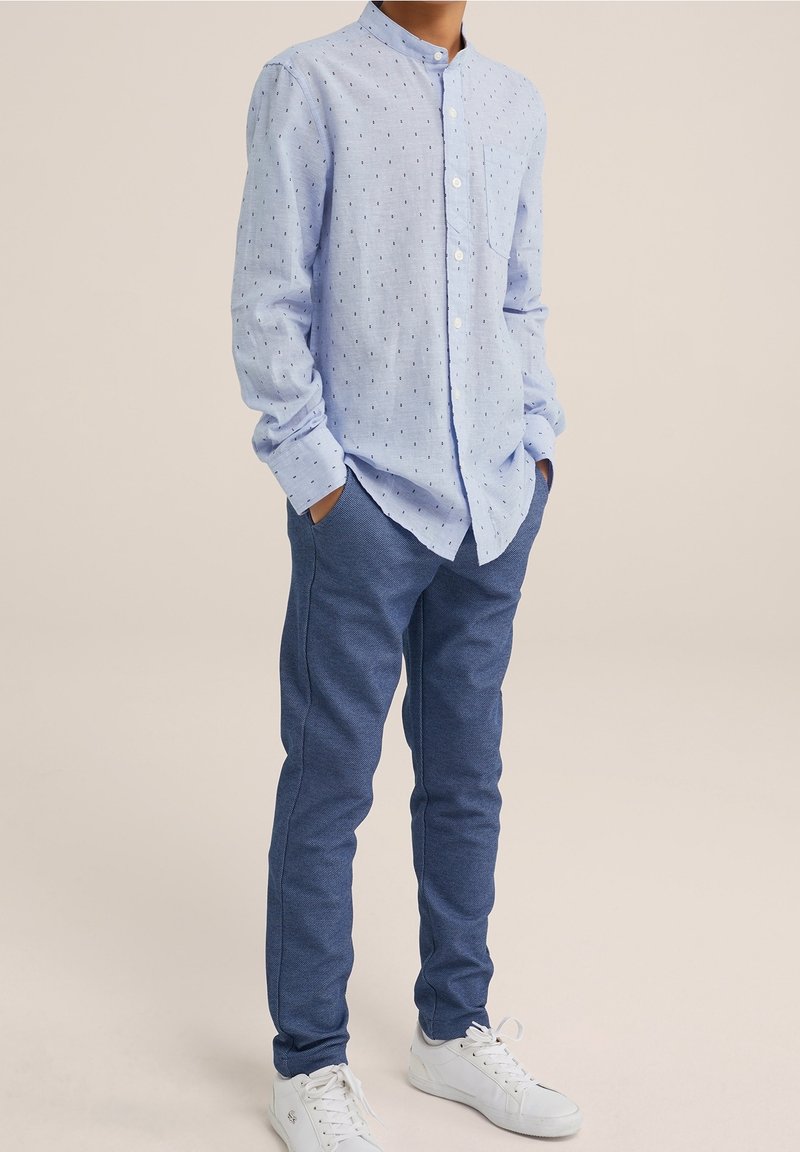 Ragazzo che indossa una camicia button-up a motivi blu chiaro, pantaloni blu, sneakers bianche, in piedi con le mani in tasca contro uno sfondo semplice.