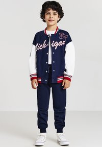 Junge mit lockigem, dunklem Haar, der eine marineblaue und weiße Collegejacke, marineblaue Jogginghosen, ein weißes T-Shirt und weiße Sneaker trägt und vor einem schlichten Hintergrund steht.