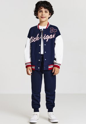 Junge mit lockigem, dunklem Haar, der eine marineblaue und weiße Collegejacke, marineblaue Jogginghosen, ein weißes T-Shirt und weiße Sneaker trägt und vor einem schlichten Hintergrund steht.