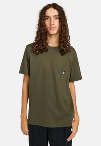 Olive green cotton T-shirt with a rounded neckline, short sleeves, and a front pocket featuring a black logo patch. Smooth texture.