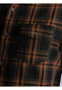 Nahaufnahme eines karierten Hemdes in Schwarz, Orange und Grün mit geknöpftem Brusttaschen und sichtbaren Knöpfen entlang der vorderen Knopfleiste.