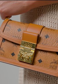 Hånd holder en tan MCM læderhåndtaske med gylden lås og sort logo-print, mod en struktureret beige strikket baggrund.