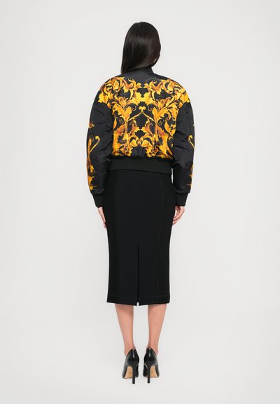 Veste bomber noire avec un brodé floral jaune vif dans le dos, associée à une jupe noire ajustée et des talons noirs, mettant en valeur une silhouette élancée.