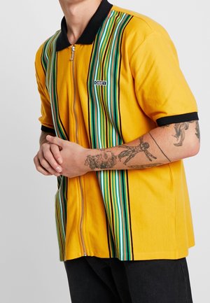 Keltainen lyhythihainen paita, jossa musta kaulus, etuvetoketju ja pystysuorat moniväriset raidat. Rinnan kohdalla "OBEY"-logo.