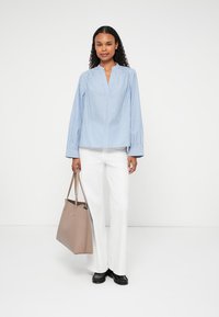 Lyseblå, stribet langærmet skjorte med v-hals, parret med hvide bukser. Modellen holder en beige tote bag og har sorte sko på.