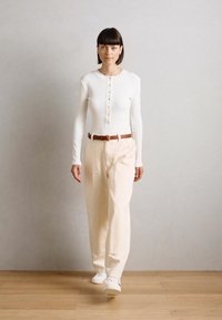 Witte geribbelde lange mouw top met knoopsluiting, gecombineerd met lichtbeige wijde broek, een bruine riem en witte sneakers op een houten vloer.