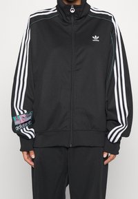 Czarna bluza sportowa na zamek wykonana z tkaniny, z białymi paskami wzdłuż rękawów i naszywką z logo w kolorowych akcentach na ramieniu.