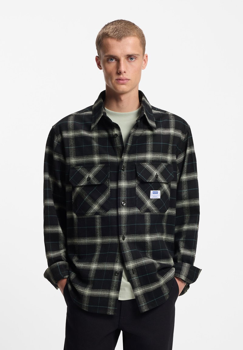 Μαύρο και πράσινο καρό πουκάμισο φανέλα με δύο τσέπες στο στήθος, κλείσιμο με κουμπιά και μια μικρή ετικέτα μάρκας στην αριστερή τσέπη.