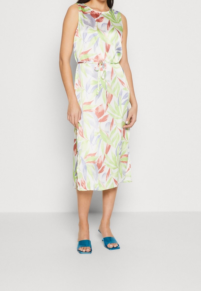 Robe sans manches à fleurs avec des motifs de feuilles vertes, rouges et violettes sur un fond blanc, dotée d'une taille coulissée. Portée avec des sandales bleues.