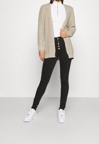 Beige knit cardigan with open front, long sleeves; paired with high-waisted black jeans featuring metal button accents and white sneakers.