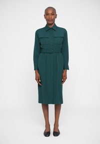 Pikkade varrukatega tume roheline kleit, millel on nööbistatud krae, kaks rinna taskut, vöödiga talje ja sirge lõige kuni põlveni.