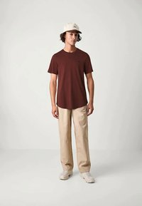 T-shirt marrone a girocollo, maniche corte, forma rilassata, abbinato a pantaloni ampi beige. Il modello indossa un cappellino chiaro e sneakers.