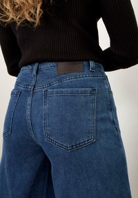 Jeans in denim blu con vita alta, dotati di tasche posteriori e di un'etichetta in pelle marrone sulla vita. Parte di una maglia nera a coste è parzialmente visibile.
