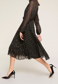 Czarna sukienka z długimi rękawami, zdobiona złotymi skośnymi paskami i lejącym dołem. W zestawie z czarnymi butami na wysokim obcasie z złotymi akcentami.