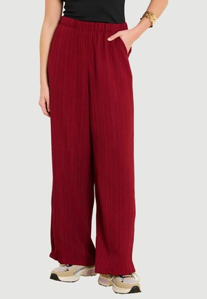 Žena oblečená v červených textúrovaných nohaviciach s rozšírenými nohavicami a elastickým pásom, čiernom topu, zlatom náramku a béžových teniskách, stojaca s jednou rukou v vrecku.