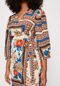 Wikkeljurk met een kleurrijk bloemen- en geometrisch patroon in crème, blauw en bruin, met driekwartmouwen en een ceintuur om de taille.