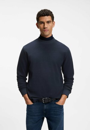 Mann mit lockigem Haar, der einen dunkelblauen Pullover und blaue Jeans trägt, steht mit den Händen in den Taschen vor einem einfarbigen weißen Hintergrund.
