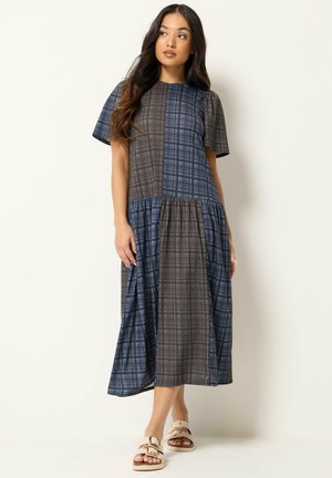 Frau trägt ein blau-graues Patchwork-Karo-Midikleid mit kurzen Ärmeln und beigen Sandalen, stehend vor einem schlichten hellen Hintergrund.