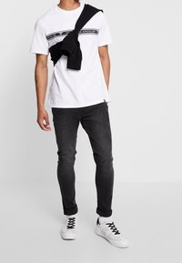 Bílý tričko s krátkým rukávem a černým textem, spárované s tmavými džíny. Černý svetr uvázaný kolem ramen. Bílé tenisky s černými tkaničkami.