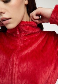 Giacca zip-up rossa in peluche con colletto alto e texture a zig-zag. La zip è decorata con gemme. Scorcio ravvicinato dell'area del collo e del tessuto morbido.