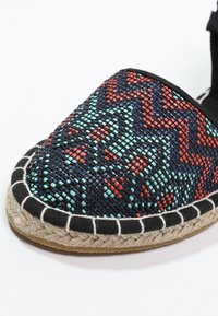 Espadrilla intrecciata con base nera, caratterizzata da un motivo geometrico rosso e blu, suola in juta e accenti in tessuto nero lungo il bordo.