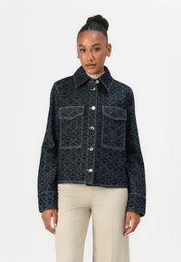Svart denimjakke med geometrisk blått mønster, spiss krage, to brystlommer og sølvfargede trykknapper, brukt over en kremfarget topp.