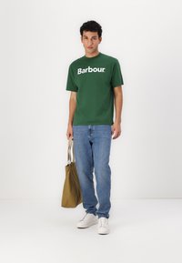 Πράσινο βαμβακερό γραφικό t-shirt με λογότυπο "Barbour", συνδυασμένο με μπλε τζιν και λευκά αθλητικά παπούτσια. Το μοντέλο κρατά μια καφέ τσάντα.