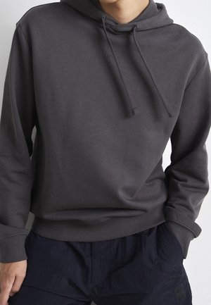 Tummanharmaa huppari, jossa on kiristysnauhat, pidetty mustujen housujen kanssa, kädet osittain taskuissa, vaatteissa näkyvät "HUGO" ja "BOSS" -logot.