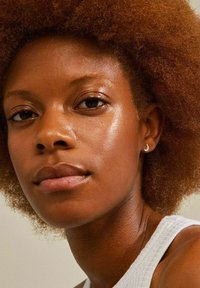 Close-up van een persoon met gekruld, kastanjebruin afro haar, die een kleine zilveren ringoorbel draagt, met gladde, stralende huid en een top in neutrale tinten.