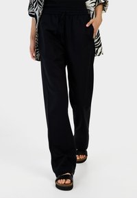 Zwarte, loszittende broek met een elastische tailleband en trekkoord, gemaakt van lichte stof. Gecombineerd met zwarte platte sandalen.